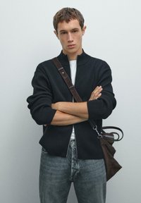 Un giovane con i capelli corti e castani sta in piedi con le braccia incrociate, indossando un maglione nero con zip, jeans grigi e una borsa a tracolla marrone.