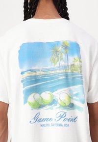 Hvid T-shirt med et blåt og grønt grafisk design af en tennisbane, palmetræer og tennisbolde med teksten "Game Point Malibu" nedenunder.