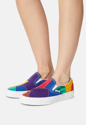 Des chaussures slip-on colorées en daim, avec un design en patchwork comprenant des sections en violet, rouge, bleu, vert et jaune. Semelle en caoutchouc blanche.