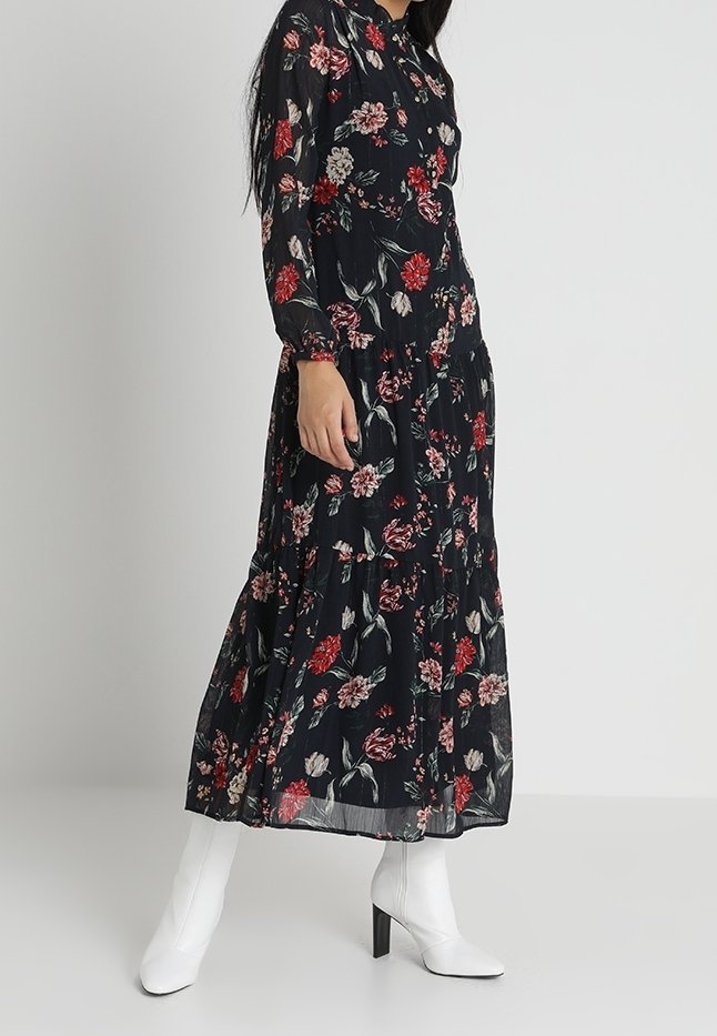 Femme portant une longue robe noire à fleurs rouges et roses, associée à des bottines blanches à talons, debout devant un fond gris uni.