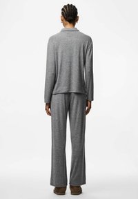 Persona ar pīņiem matos, ģērbusies pelēkā garā piedurknē augšpusē un biksēs, stāv ar muguru pret skatītāju, ar brūnām kurpēm uz baltā fona.