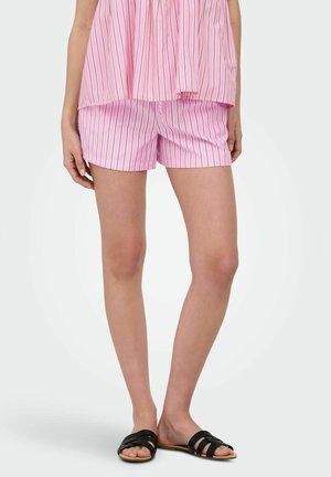 Kvinde iført pink og hvide lodrette stribede shorts og matchende top, stående barfodet med sorte slides mod en hvid baggrund.