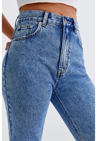 Jean en denim bleu taille haute avec un délavage, design à cinq poches et coutures contrastées, mettant en avant un tissu lisse et texturé.