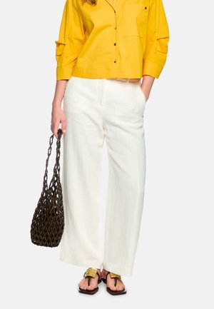 Woman wearing yellow button-up top, white wide-leg pants, gold sandals, holding large dark brown woven bag by side.