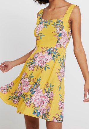 Robe jaune sans manches avec des motifs floraux roses et des feuilles vertes, portée par une personne aux cheveux bouclés, en mouvement montrant le déplacement de la robe.