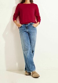 En person som bär en röd långärmad tröja, ljusblå raka jeans och bruna slip-on skor, står med händerna i fickorna mot en vit vägg.