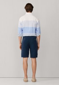 Hombre de pie de espaldas, con camisa a rayas blancas y azul claro, pantalones cortos azul marino y zapatos beige sin cordones.