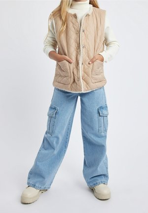 Dziecko ubrane w beżową pikowaną kamizelkę na kremowym swetrze, jasnobłękitne jeansy typu cargo oraz kremowe buty, stojące z rękami w kieszeniach kamizelki.