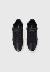 Adidași atletici negri, din piele și textil, având o talpă albă, șireturi negre și un logo pe limbă.
