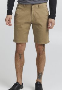 Shorts en coton beige avec une coupe droite, passants de ceinture et poches avant. Présentent un ourlet cousu et des tatouages visibles sur les jambes inférieures.