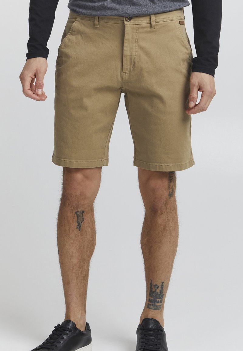 Shorts en coton beige avec une coupe droite, passants de ceinture et poches avant. Présentent un ourlet cousu et des tatouages visibles sur les jambes inférieures.