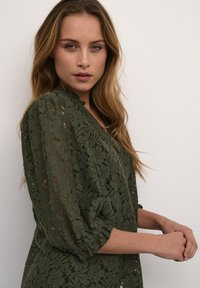 Oliven grøn blonde top med blomstermønster, 3/4 pufærmer og en scalloped halsudskæring. Tekstureret stof med en gennemsigtig kvalitet.
