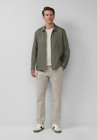 Olijfgroene linnen shirt met een kraag en een borstzak, gecombineerd met lichtbeige broek en witte sneakers met zwarte accenten.