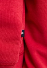 Tessuto di felpa rossa con una texture a coste, con un piccolo etichetta blu navy con logo bianco sul lato. Cuciture visibili sull'orlo e sui polsini.