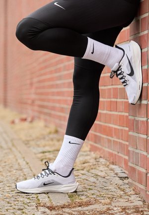 Witte Nike hardloopschoenen met zwarte accenten, een bovenwerk van mesh, een grijze zool, gecombineerd met witte sokken met een zwart logo, gedragen bij zwarte legging.