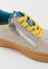 Sneaker with a beige suede upper, textured metallic accents, yellow laces, blue tongue, and side zipper. Rubber sole in natural color.