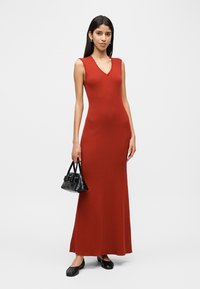 Γυναίκα που φοράει αμάνικο, μακρύ μέχρι το πάτωμα φόρεμα σε σκουρόκόκκινο χρώμα, κρατώντας μια μικρή μαύρη τσάντα χειρός και μαύρα μπαλαρίνες, σε άσπρο φόντο.