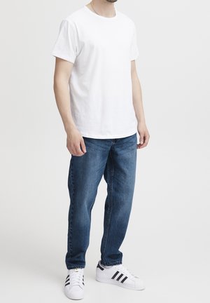 Homme portant un t-shirt blanc uni à manches courtes, un jean bleu et des baskets blanches à rayures noires, debout devant un fond clair uni.