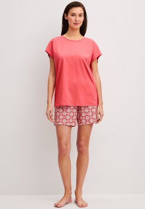 Frau steht barfuß, trägt ein korallenrotes kurzärmeliges Oberteil und gemusterte rote und weiße Shorts vor einem schlichten hellen Hintergrund.