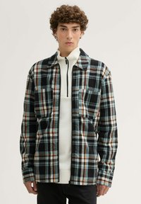 Karierter Flanelljacke in Schwarz, Blau und Orange, mit zwei Brusttaschen, Frontreißverschluss und Kragen. Über einem weißen Pullover getragen.