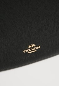 Surface en cuir noir texturé avec un petit logo Coach doré et un emblème de carriole au centre en bas.