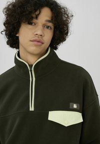 Pull polaire vert foncé avec une demi-fermeture éclair, un col vert clair et une poche poitrine. Présente un logo sur la poche et une texture douce.