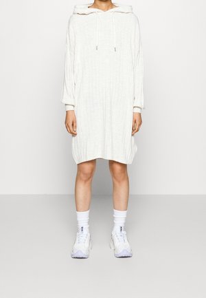 Personne portant une robe à capuche en tricot blanc à manches longues, assortie à des chaussettes Nike blanches et des baskets blanches, se tenant sur un fond uni.