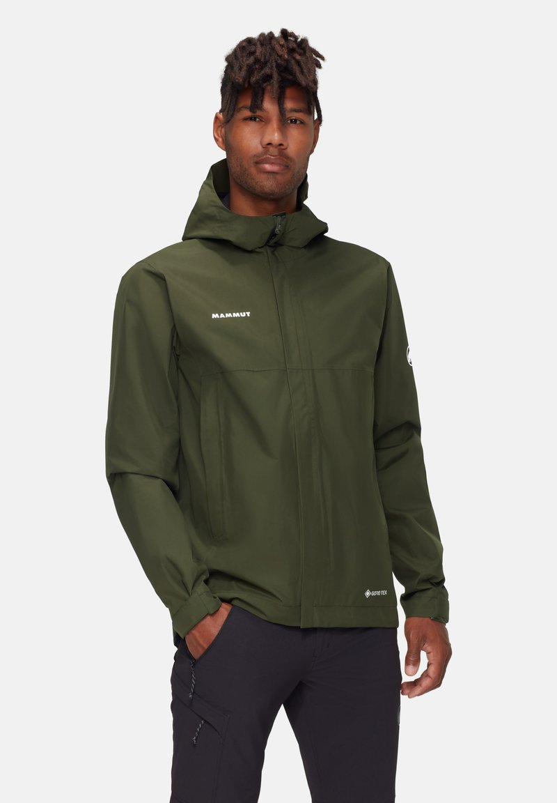 Giacca impermeabile verde oliva con cappuccio, chiusura con zip, due tasche laterali e logo "Mammut" sul petto. Presenta il marchio Gore-Tex.