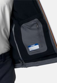 Wnętrze czarnej polarowej kurtki z szarym zamkniętym kieszonką. Kieszeń posiada niebieską etykietę na imię i dane. Zamek jest srebrny.