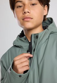 Light green waterproof jacket with a hood, zippered front, and snap buttons. Material appears smooth with a matte finish and subtle lining.