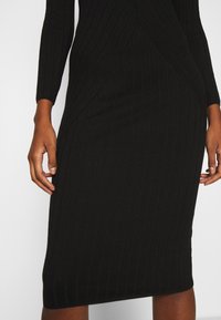 Robe noire en tricot côtelé avec des manches longues, silhouette ajustée et texture à rayures verticales. Les détails des coutures soulignent la forme de la robe.