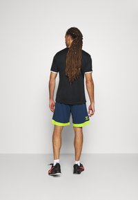 Svart idrottströja med vit kant, kombinerad med marinblå shorts med neongul fåll. Bärs med svarta sneakers, sedd bakifrån.