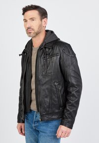 Zwarte leren jas met een grijze capuchon, voorzien van meerdere ritszakken en een gladde textuur. Draag een beige shirt en blauwe spijkerbroek.