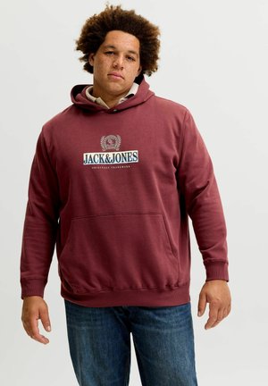 Jeune homme aux cheveux bouclés portant un sweat à capuche bordeaux Jack & Jones et un jean bleu, debout devant un fond uni clair.