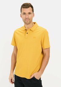 Koszulka polo z krótkim rękawem w kolorze musztardowym, wykonana z teksturowanej tkaniny. Posiada klasyczny kołnierzyk, zapięcie na dwa guziki oraz naszywkę z logotypem.