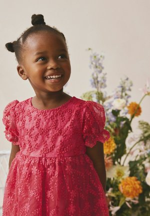 Smilende ung pige med to hårdutter og iført en lyserød broderet kjole står foran farverige blomster.