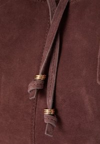 Καφέ σουέντ ύφασμα με λεία υφή, με διακοσμητική λεπτομέρεια κόμπου και λεπτομέρειες από χρυσό μέταλλο στα δαχτυλίδια του κορδονιού.