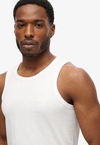 Camiseta sin mangas blanca de algodón con cuello redondo, que presenta un pequeño logo bordado en el pecho. La tela parece suave y ligera.