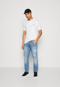 Hvit polo skjorte med korte ermer og en liten logo, kombinert med lyseblå ribbede jeans og svarte joggesko, komplementert av en svart caps.