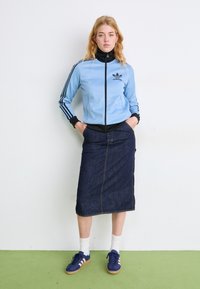 Casaco azul claro com fecho de correr, detalhes em preto e três riscas, combinado com saia comprida de ganga, meias brancas e ténis azuis.