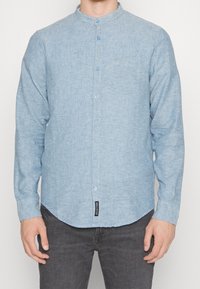 Chemise bleu clair à boutons, en tissu léger, col rond, manches longues, poche poitrine unique, ourlet arrondi, étiquette en bas à gauche.