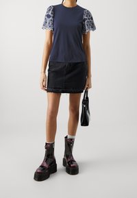 Kvinna som bär en marinblå topp med mönstrade spetsärmar, svart denimkjol, plattform stövlar, och håller en svart handväska mot en enfärgad bakgrund.