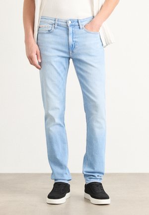 Jeans en denim bleu clair avec une coupe slim, cinq poches et un délavage. Associés avec des baskets noires et une chemise blanche.