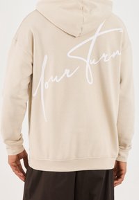 Osoba w beżowej bluzie z kapturem, z białym kursywą napisem "Twój ruch" na plecach, w zestawieniu z ciemnymi spodniami.