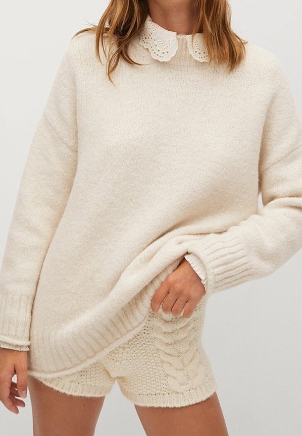 Femme portant un pull en maille crème avec un col en dentelle et un short assorti en maille torsadée, tenant l'ourlet du pull d'une main.