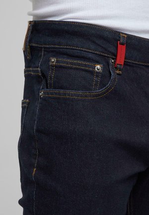 Jean en denim foncé avec des coutures jaunes, comportant une poche avant, une fermeture à bouton et une étiquette de marque rouge sur la taille.