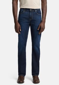 Dunkelblaue Jeans für Herren mit geradem Schnitt, mittelhoher Taille, dezenten Nähten und Fünf-Taschen-Design. Getragen mit braunen Schuhen.