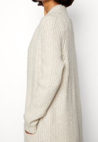 Cremfarbener, lockerer Strickcardigan mit gerippter Textur, langen Ärmeln und seitlichen Taschen.