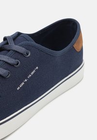 Marineblaue Segeltuchschuhe mit grauen Schnürsenkeln, weißer Gummi-Sohle und braunem Lederakzent an der Ferse. Zeigt geografische Koordinaten an der Seite.