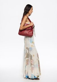 Rdeča usnjena torba čez ramo z teksturiranim zaključkom, črn trak in srebrni dodatki. Model nosi lahkotno, vzorčasto maxi krilo z prosojnim pokrivalom.